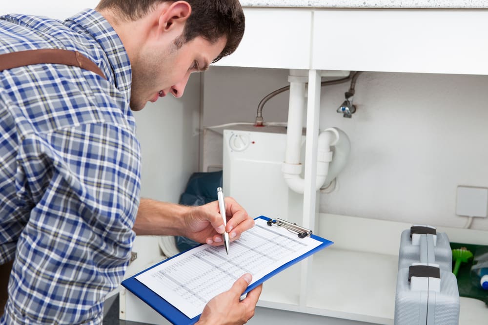 Plumber inspecting a pipe under a sink with a clipboard.