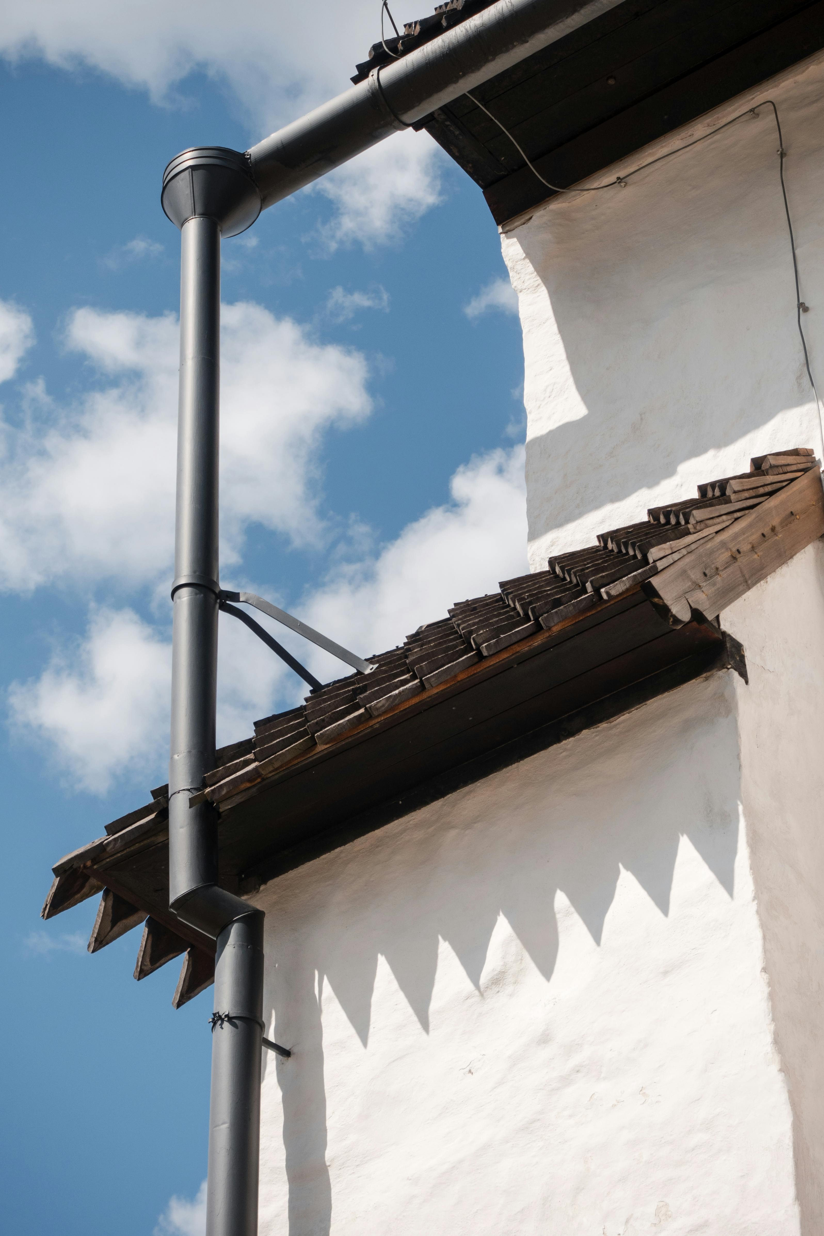 Close-up of a building's roofline with a metal pipe and white wall against a blue sky.