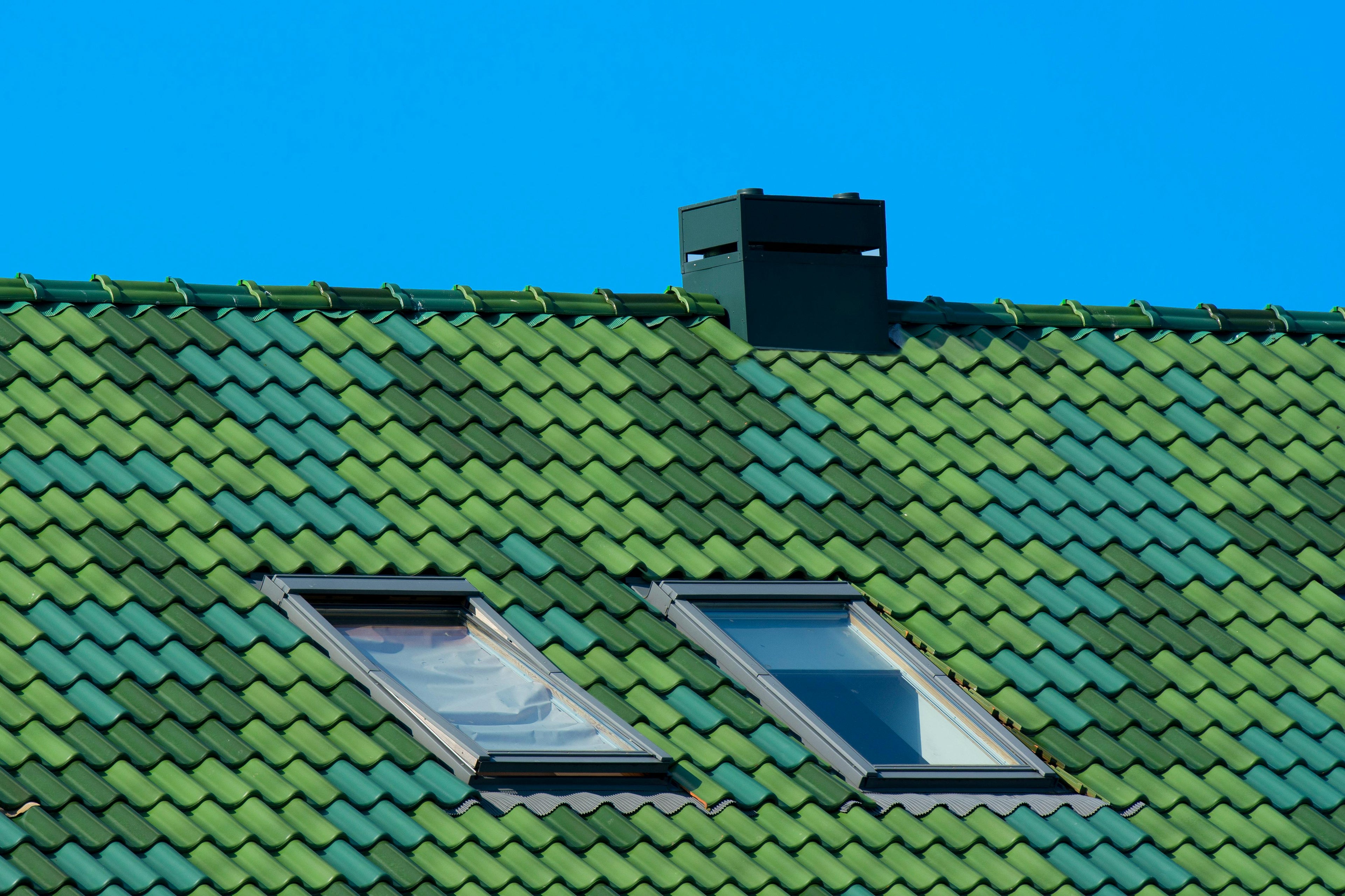 Green tiled roof with two skylights and a chimney against a clear blue sky.