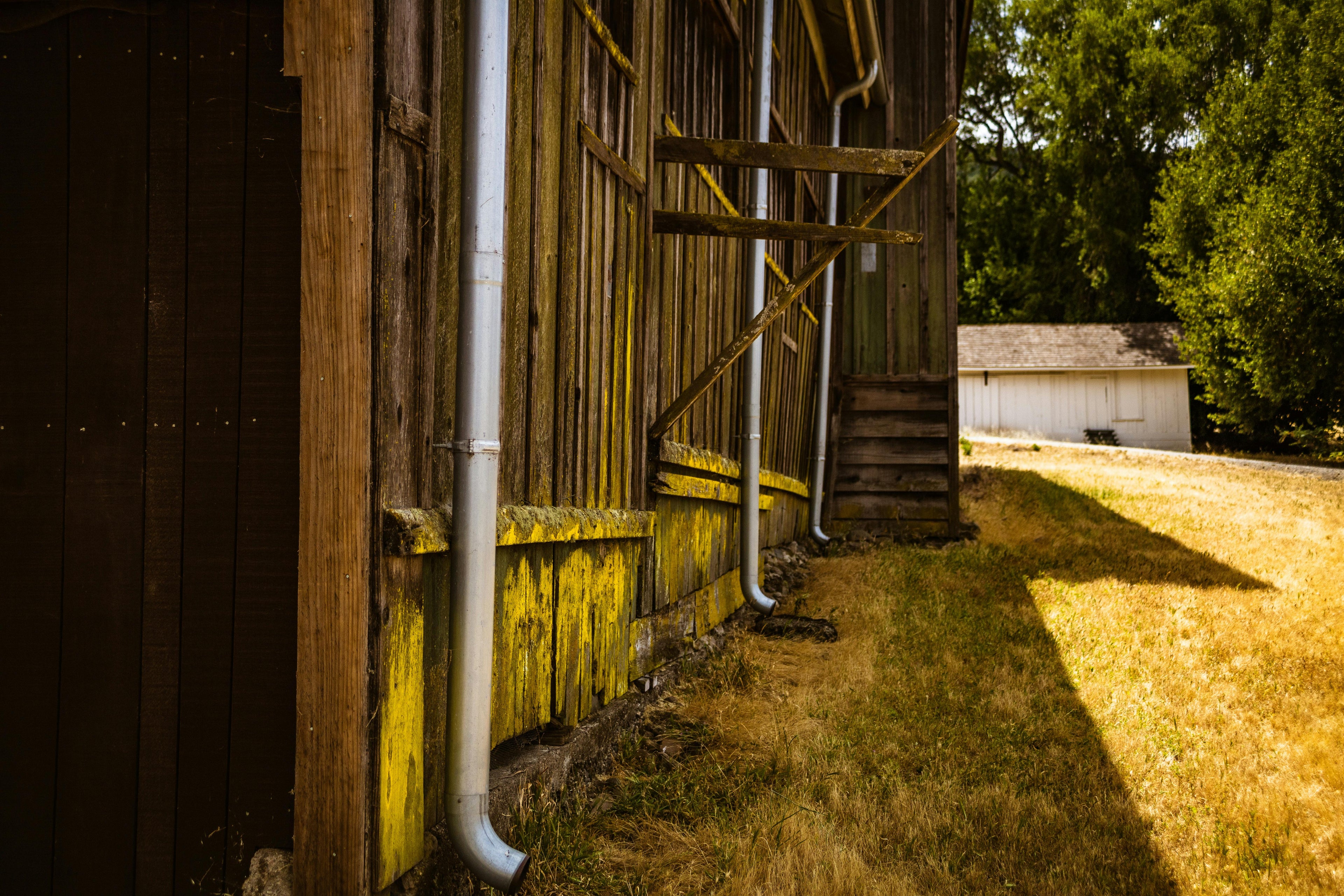 Old wooden building with a white pipe and staircase, surrounded by greenery.